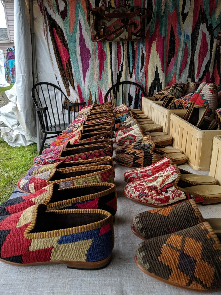 Rows of kilim shoes lined up for sale on a table, with a Moroccan rug hanging in the background.