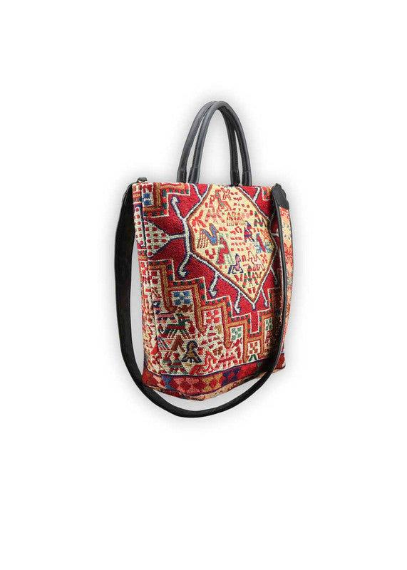 The Artemis Design Co. Sumak Kilim Tote is a stylish and versatile accessory crafted with a blend of sumptuous brown leather and vibrant, eye-catching colors. The tote features a traditional Sumak Kilim pattern in a spectrum of hues including red, white, blue, green, peach, orange, and yellow. This eclectic color combination lends a dynamic and playful aesthetic to the bag, making it a statement piece for any outfit or occasion.  (Side View)