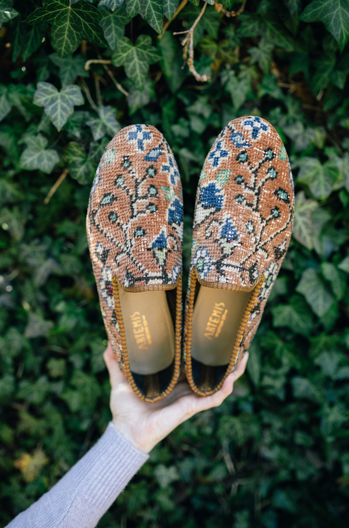 woman holding men's carpet loafers in front of an ivy covered wall