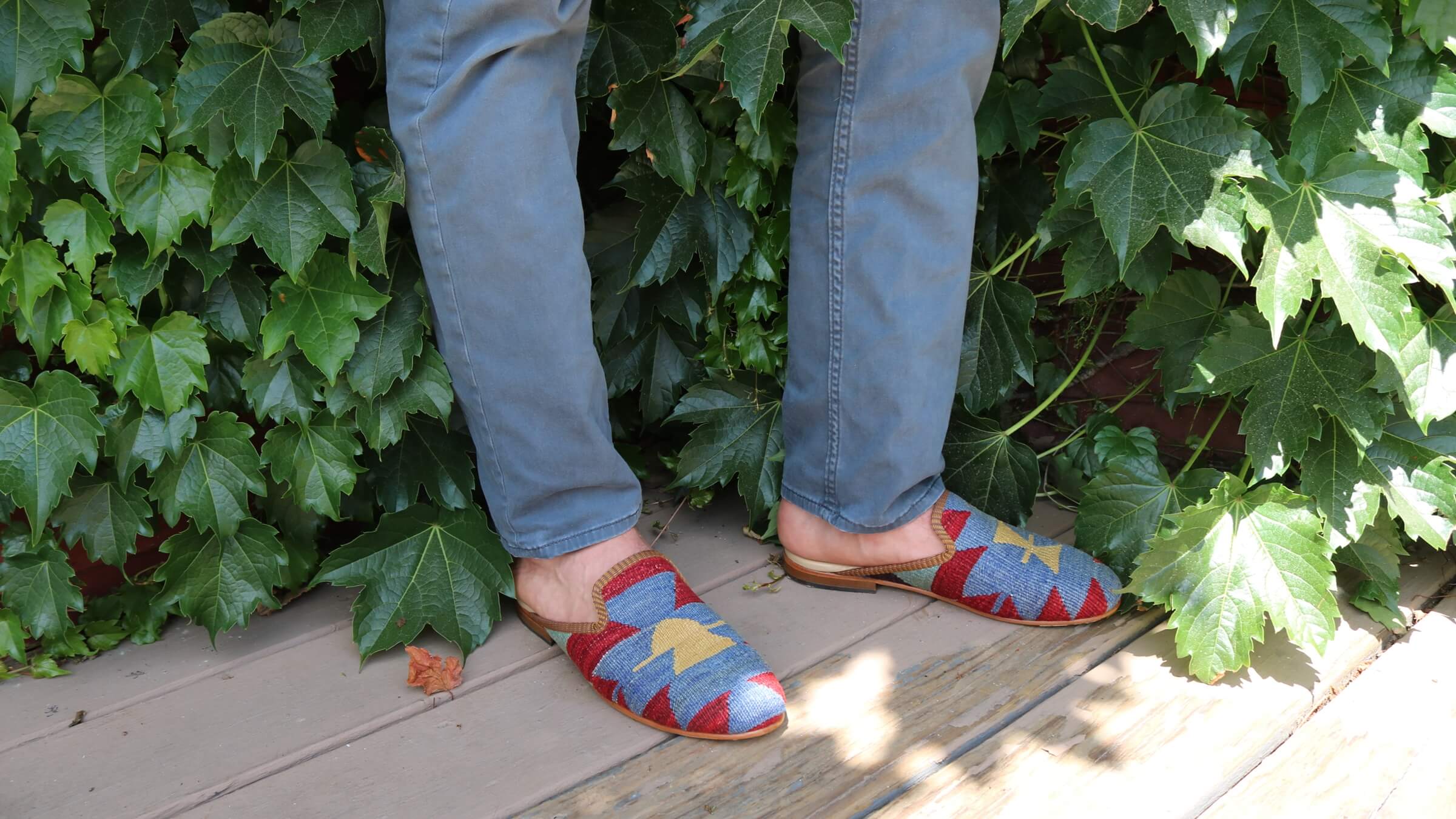 Person wearing blue jeans and colorful sandals standing on a wooden deck with green foliage in the background.