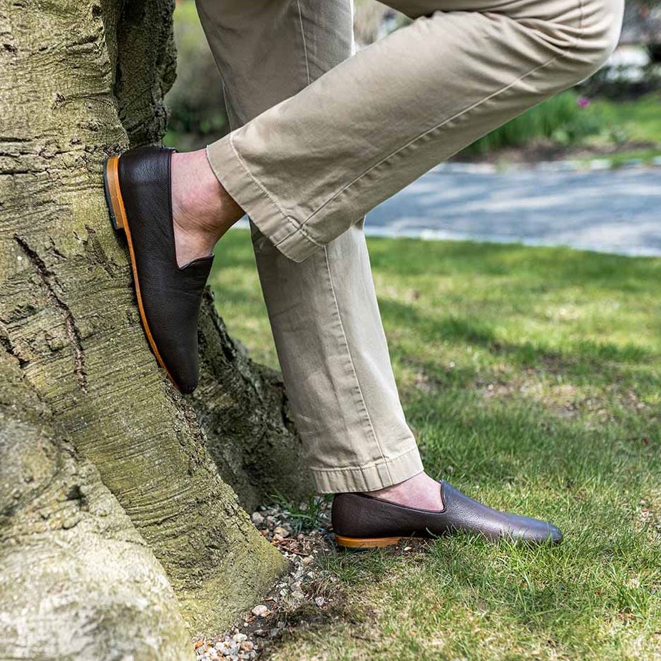 A man's legs wearing khaki pants and brown leather Artemis Design Co. loafers, leaning against a tree.