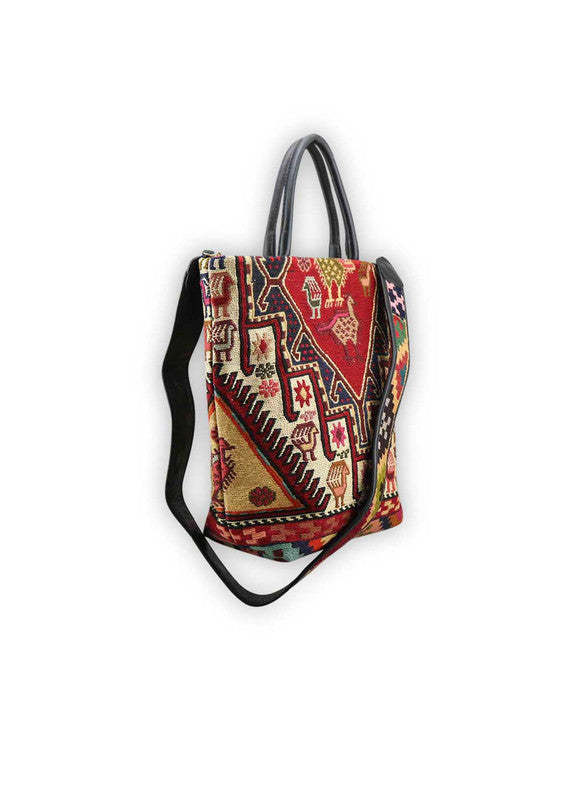 The Artemis Design Co. Sumak Kilim Tote is a stylish and versatile accessory crafted with a blend of sumptuous brown leather and vibrant, eye-catching colors. The tote features a traditional Sumak Kilim pattern in a spectrum of hues including red, white, blue, green, peach, orange, and yellow. This eclectic color combination lends a dynamic and playful aesthetic to the bag, making it a statement piece for any outfit or occasion. (Side View)