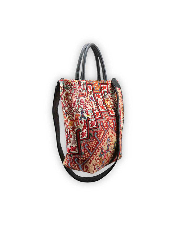 The Artemis Design Co. Sumak Kilim Tote is a stylish and versatile accessory crafted with a blend of sumptuous brown leather and vibrant, eye-catching colors. The tote features a traditional Sumak Kilim pattern in a spectrum of hues including red, white, blue, green, peach, orange, and yellow. This eclectic color combination lends a dynamic and playful aesthetic to the bag, making it a statement piece for any outfit or occasion. (Side View)
